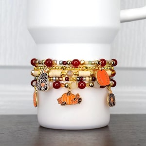 May include: A stack of beaded bracelets featuring gold, red, and cream-colored beads. The bracelets include several metal charms shaped like pumpkins, some with orange enamel and others with black and white floral patterns.
