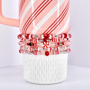 Puede incluir: Un conjunto de pulseras de cuentas rojas, blancas y transparentes. Las pulseras están adornadas con diversas cuentas, incluyendo cuentas a rayas de bastón de caramelo y transparentes. Las pulseras están sobre una taza de cerámica blanca, con una taza a rayas rosas y blancas.