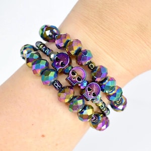 May include: Three iridescent beaded bracelets with skull charms. The bracelets feature faceted beads in shades of purple, green, and gold. Each bracelet has a small, purple skull charm. The bracelets are stacked on top of each other.