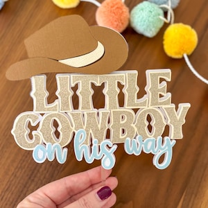 May include: A brown cowboy hat and a cake topper that reads "Little Cowboy on his way." The text is gold glitter with a light blue outline. The cake topper is held by a clear acrylic stick. Colorful pom-poms are in the background.