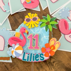 May include: A pink and blue cake topper with a flamingo, palm tree, sunglasses, and the number 11. The topper also includes the name "Lillies" in teal blue letters.