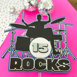 May include: A black silhouette of a drummer playing a drum set with a white circle with the number 15 in blue and the word "ROCKS" in black with glitter.