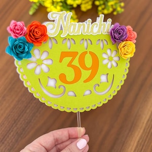 May include: A lime green cake topper with the word "Nanichi" in gold glitter and the number "39" in orange. The topper is decorated with colorful paper flowers in shades of pink, orange, teal, purple, and yellow. The topper is held by a clear plastic stick.