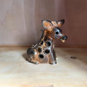 May include: A ceramic figurine of a giraffe with a glossy, mottled brown and black glaze. The body features several circular cutouts throughout its neck and torso, and the head is turned slightly to the right with pointed ears.
