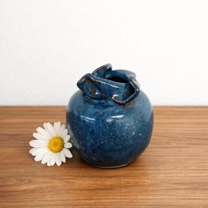 Puede incluir: Un pequeño jarrón redondo de cerámica azul con un diseño de asa único. El jarrón tiene una textura moteada y está sobre una superficie de madera junto a una margarita blanca. El jarrón mide aproximadamente 10 cm de alto.