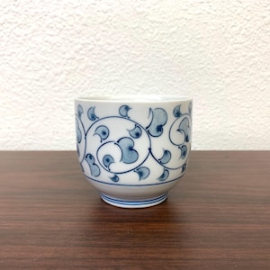 May include: A small, white ceramic teacup with a blue floral design. The cup features a repeating pattern of stylized leaves and swirls. The design is in a classic blue and white color scheme, with a simple, elegant aesthetic.