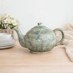 Op de afbeelding: Een keramische theepot met een gespikkeld blauw en crème design. De theepot heeft een gebogen tuit, een ronde body en een handvat. Het staat op een lichtgekleurd houten oppervlak.