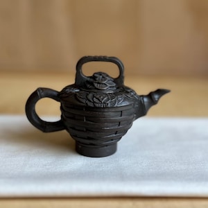 May include: A dark brown clay teapot featuring a textured exterior that mimics a woven basket pattern. The teapot includes a bamboo-shaped handle, a curved spout, and a lid adorned with carved floral details and a rectangular top handle.
