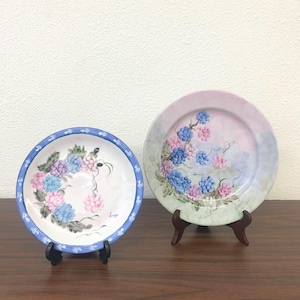 May include: Two decorative plates with floral designs. One plate has a blue border and a floral wreath of blue and pink flowers. The other plate features a similar floral arrangement in blue and pink hues, set against a pastel background.