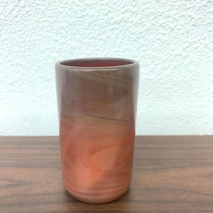 May include: A cylindrical glass with a swirling pattern of brown and peach hues. The glass has a smooth, polished finish and a rounded base. The interior of the glass is a darker shade of brown.