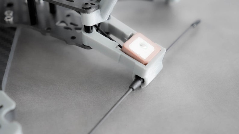 May include: Close-up of a gray drone with a small, square, white and pink antenna attached to the bottom.