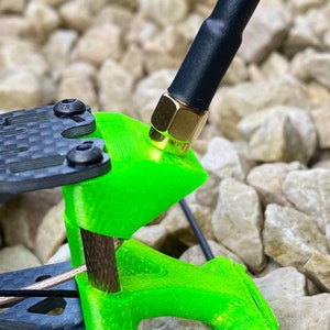 May include: A close-up of a green 3D printed antenna mount with a black antenna attached. The antenna has a gold connector. The mount is attached to a black carbon fiber frame.