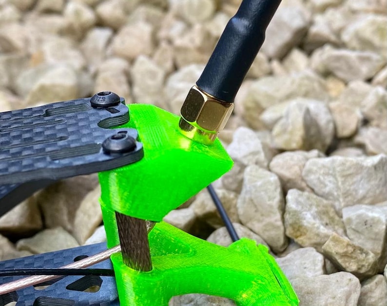 May include: A close-up of a green 3D printed antenna mount with a black antenna attached. The antenna has a gold connector. The mount is attached to a black carbon fiber frame.