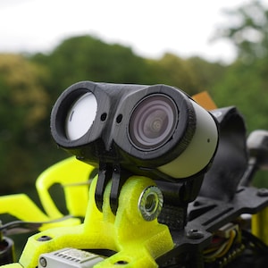 May include: A black and white dual-lens camera mounted on a yellow 3D printed mount. The camera has a black lens cover on the left side and a clear lens cover on the right side.