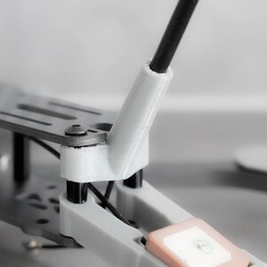 May include: Close-up of a drone frame with a white plastic antenna mount and a black antenna. The frame is made of metal and has a gray finish.