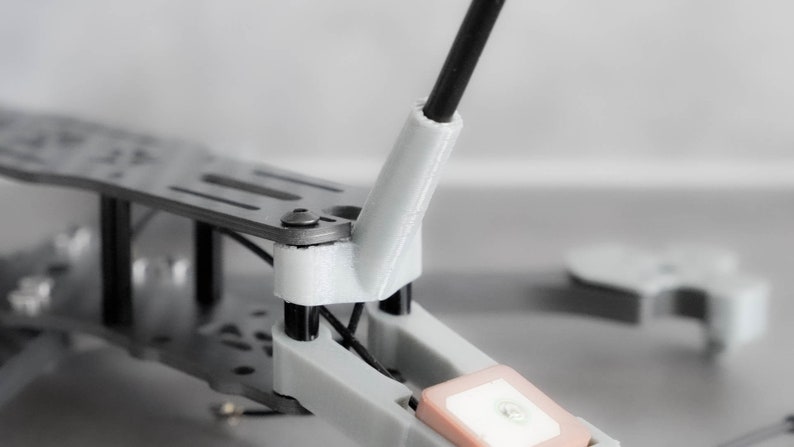 May include: Close-up of a drone frame with a white plastic antenna mount and a black antenna. The frame is made of metal and has a gray finish.