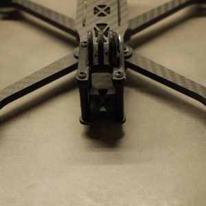 May include: A black carbon fiber drone frame with gray plastic landing gear. The frame has a cross shape with four arms and a center section. The landing gear is attached to the ends of the arms.