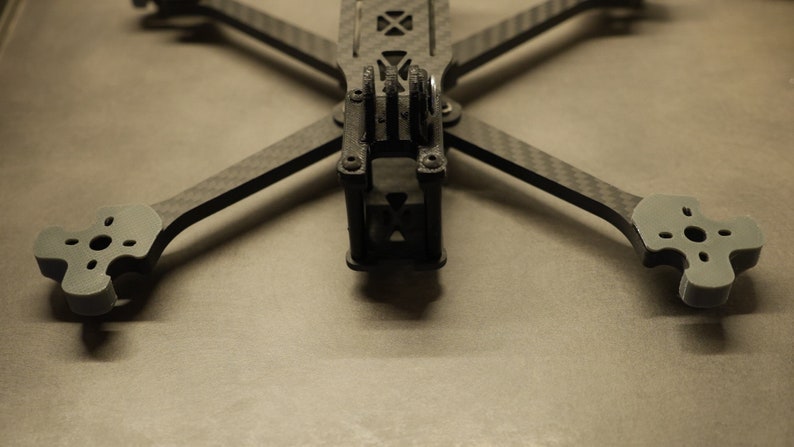 May include: A black carbon fiber drone frame with gray plastic landing gear. The frame has a cross shape with four arms and a center section. The landing gear is attached to the ends of the arms.