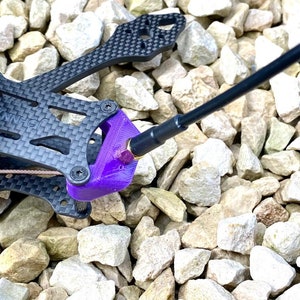 May include: A close-up of a purple 3D printed drone frame stand with a black carbon fiber drone frame and a black screwdriver. The stand is designed to hold the drone frame in place while working on it.