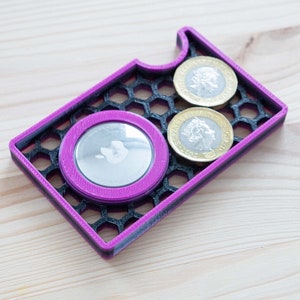 May include: A pink and black 3D printed card holder with a honeycomb pattern. The holder has a slot for a card and two slots for coins. Two gold coins are visible in the slots.