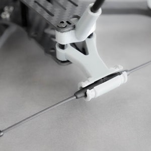 May include: A close-up of a white drone frame with a black antenna attached to the front. The antenna is secured with a white plastic piece.