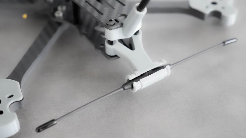 May include: A close-up of a white drone frame with a black antenna attached to the front. The antenna is secured with a white plastic piece.