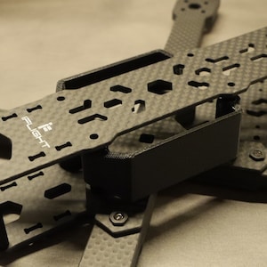 Op de afbeelding: Een zwart carbon fiber droneframe met de tekst "iflight" erop gedrukt. Het frame is ontworpen voor een quadcopter drone en heeft een zwarte plastic doos in het midden.