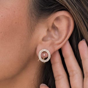 May include: A circular stud earring featuring a gold-toned metal frame encrusted with small, clear, sparkling stones arranged in a pave setting. The design consists of a hollow ring shape with a central horizontal bar, creating a geometric aesthetic.