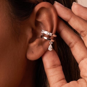 May include: A silver-toned ear cuff featuring two stacked bands. The upper band is set with rectangular clear stones, while the lower band includes a dangling teardrop-shaped clear stone charm that hangs from the bottom edge of the metal.
