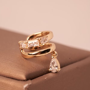 May include: A gold-toned ear cuff featuring a double-band design. The upper band is embellished with three clear, faceted stones, including a central baguette cut. A single pear-shaped clear stone hangs from the lower band as a dangling charm.