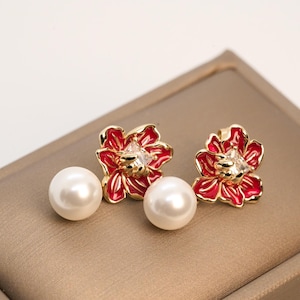 May include: A pair of floral stud earrings. Each earring features a red flower with gold trim and a central clear gem. A large, round, white pearl sits below each flower. The earrings are displayed on a gold-toned surface.
