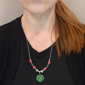 May include: A silver chain necklace with a green frog pendant. The necklace features red and white polka dot beads, green beads, and a green frog pendant. The necklace is worn over a black shirt and cardigan.