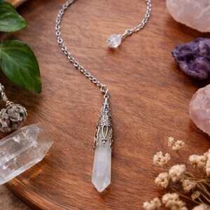 May include: A clear quartz crystal pendulum with a silver chain and decorative cap. The pendulum is displayed on a wooden tray with other crystals and dried flowers. The background is a warm, natural wood tone.