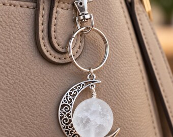 May include: A silver-toned keychain featuring a crescent moon charm with intricate filigree cutouts and a round, translucent, textured white stone sphere nestled within the curve of the moon, attached to a circular ring and a lobster claw clasp.