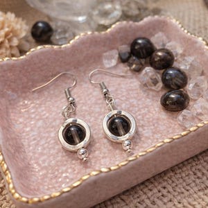 May include: A pair of silver earrings with a dark gray, round stone set in a silver circle. The earrings are displayed in a pink, scalloped dish with gold trim, surrounded by dark gray stones and clear crystals.
