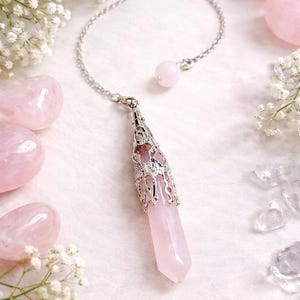 May include: A pink rose quartz crystal pendulum with a silver chain and decorative cap. The crystal is surrounded by other rose quartz stones and white flowers. The pendulum is a light pink color.