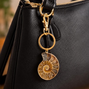 May include: A black textured handbag with a gold-toned metal keychain attached to the handle. The keychain features a polished ammonite fossil pendant with brown and tan spiral patterns, connected by a gold ring and a lobster claw clasp.