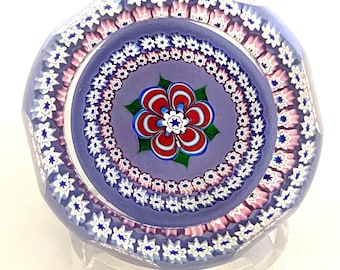 May include: A multi-layered glass paperweight with a purple background and a red, white and green flower design in the centre. The paperweight is surrounded by concentric circles of white stars and pink and white flowers.