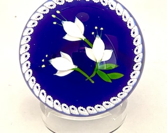 May include: A circular glass paperweight featuring a deep blue background with three white lily-like flowers and green leaves in the centre. The outer edge is decorated with a white and blue floral millefiori pattern border.