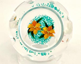 May include: A clear glass paperweight featuring a circular design with two yellow flowers and green leaves centred against a textured turquoise background. The glass has a scalloped outer edge and sits on a circular clear glass base.