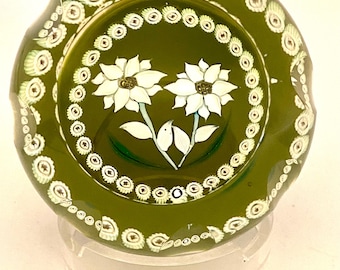 May include: A round olive green glass paperweight featuring a central design of two white flowers with green leaves, surrounded by a circular border of small white and green millefiori glass canes, with additional canes along the scalloped edge.