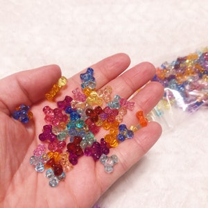 May include: A handful of colorful, translucent plastic beads in various shapes and sizes. The beads are in shades of blue, pink, yellow, orange, and purple. Some beads are scattered on a white surface, while others are in a clear plastic bag.
