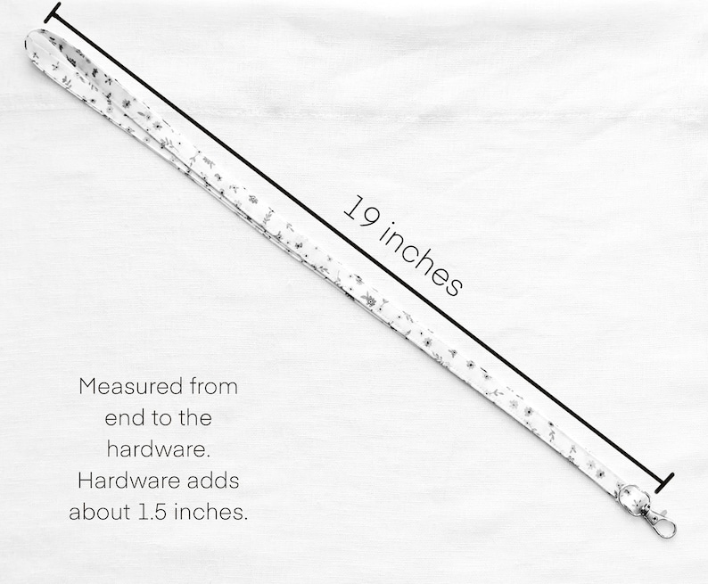 Op de afbeelding: Een wit stoffen koord met een klein zwart bloemenpatroon. Het koord meet 19 inch van het uiteinde tot de hardware, waarbij de metalen hardware ongeveer 1,5 inch toevoegt. De tekst luidt: 19 inches. Measured from end to the hardware. Hardware adds about 1.5 inches.