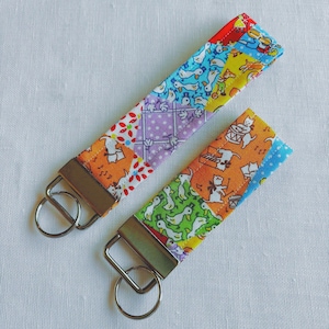 May include: Two colorful fabric key fobs with metal hardware. The key fobs feature a patchwork design with various patterns, including ducks, cats, and floral prints. The key fobs are in shades of orange, purple, green, and blue.