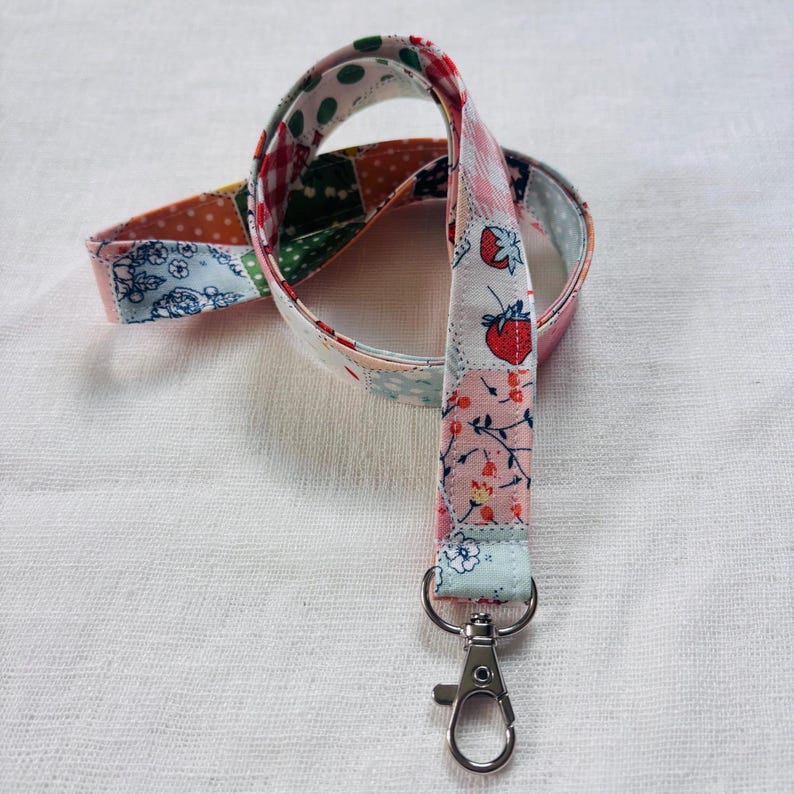 Op de afbeelding: Een stoffen lanyard met een patchworkontwerp met diverse patronen, waaronder rode aardbeien, bloemmotieven, stippen en gingham-ruitjes in tinten roze, blauw, groen en oranje, afgewerkt met een zilverkleurige metalen karabijnhaak.