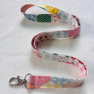 May include: A fabric lanyard featuring a patchwork design with various pastel-colored patterns, including small strawberries, green polka dots, yellow flowers, and light blue gingham checks, finished with a silver-toned metal swivel lobster clasp.