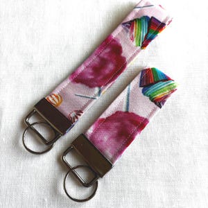 May include: Two key fobs with a pink background and a watercolor design featuring cotton candy and rainbow accents. Each fob has a metal key ring and a rectangular metal piece for attaching the fabric.