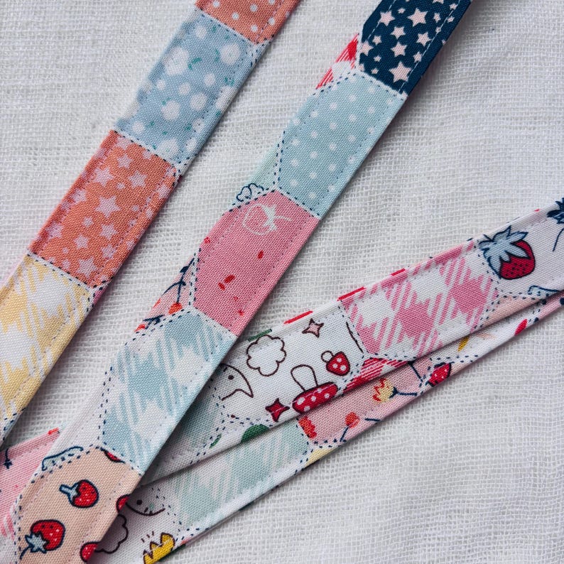 Op de afbeelding: Een stoffen band met een patchworkontwerp van diverse patronen, waaronder lichtblauwe stippen, oranje sterren, roze ruitjes en witte gedeelten met kleine rode illustraties van aardbeien, paddenstoelen en wolken. De band heeft zichtbare stiksels.