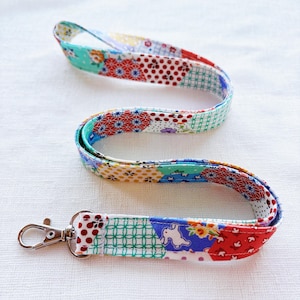 May include: A colorful fabric lanyard with a metal clasp. The lanyard is made of various patterned fabric patches in red, blue, green, and yellow. The fabric has floral, polka dot, and geometric designs. The lanyard is approximately 30 inches long.