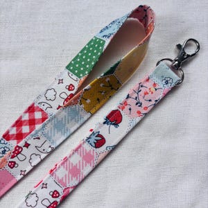 Op de afbeelding: Een stoffen lanyard met een patchworkontwerp van diverse patronen, waaronder rood-witte vichyruitjes, groene stippen, aardbeien, paddenstoelen en bloemmotieven. De band is bevestigd aan een zilverkleurige, draaibare metalen karabijnhaak.